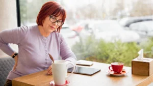 Mature woman holds her lower back at a sunlit cafe table to ease immediate stiffness from prolonged sitting.