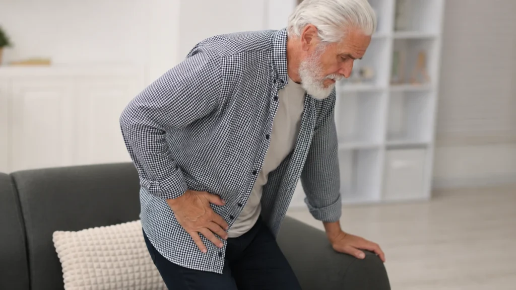 Mature man presses his front hip crease while rising from a low sofa to stretch tight muscles.