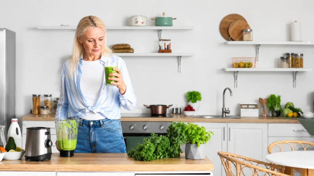 Mature woman standing in a sunlit kitchen holding a raw green kale smoothie to support her thyroid health after 50.