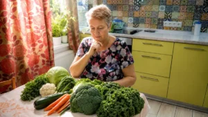 A woman in her 60s sits at a kitchen table looking pensively at a spread of cruciferous vegetables including broccoli, kale, cauliflower, and cabbage, with her hand raised to her chin in concern.