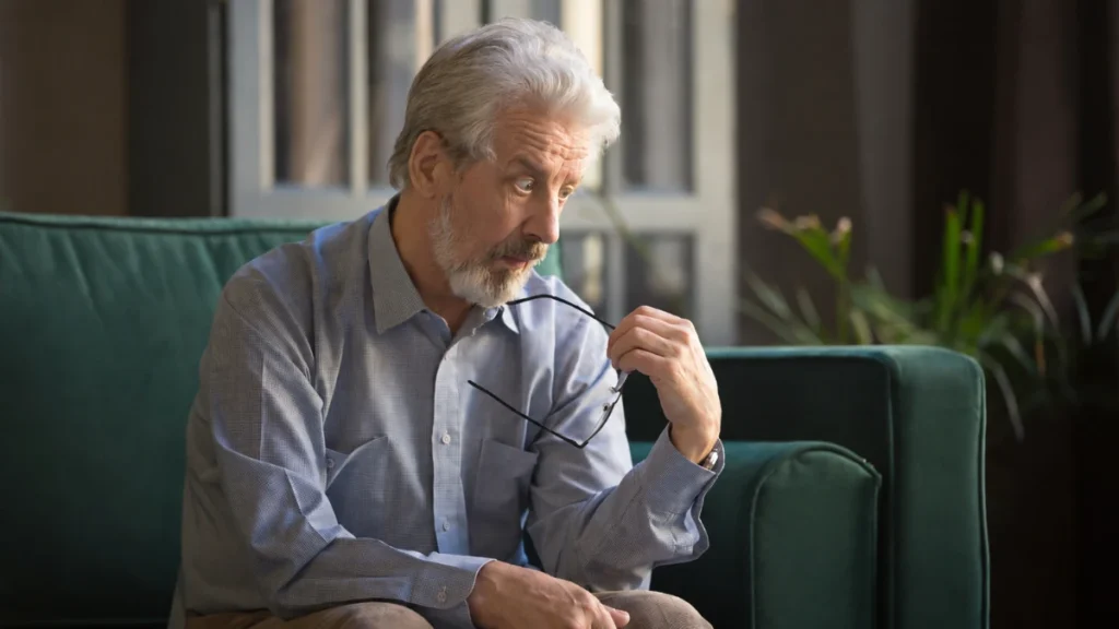 Man in his 60s seated at home, holding glasses, brow furrowed with a look of unlocatable concern and no visible cause.