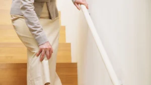 Older woman gripping a home stair railing with one hand and pressing the other to her knee mid-climb, the instinctive pause of someone familiar with knee pain on stairs.