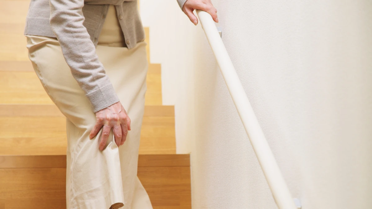 Older woman gripping a home stair railing with one hand and pressing the other to her knee mid-climb, the instinctive pause of someone familiar with knee pain on stairs.