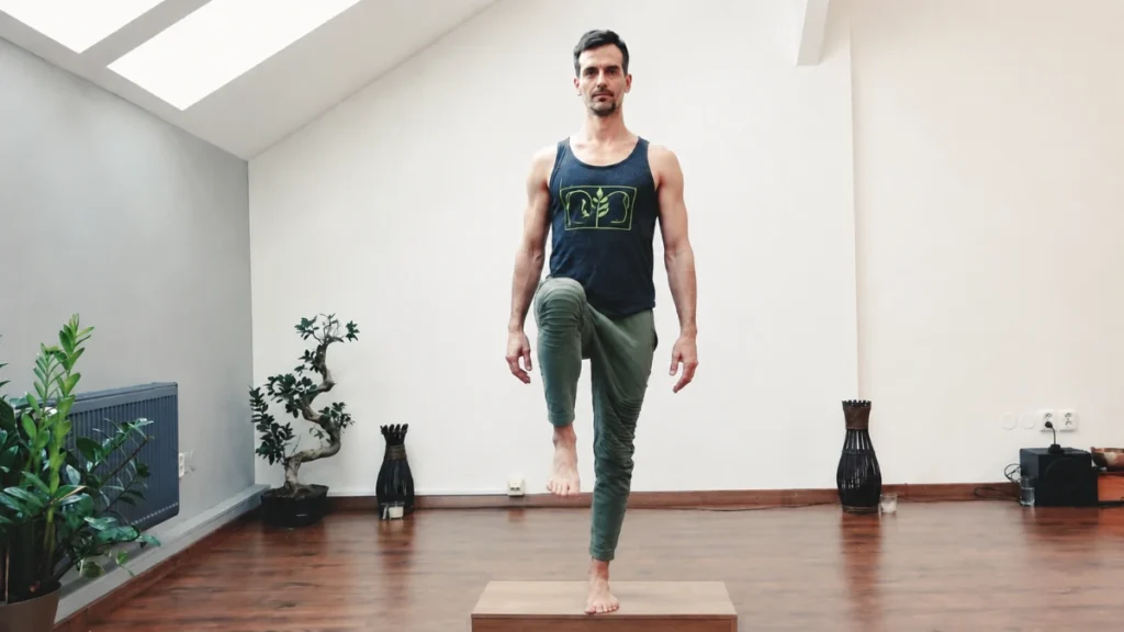 Man balancing on one leg on a low wooden step in a bright home studio, opposite knee raised, holding steady upright posture to engage hip stability and control.