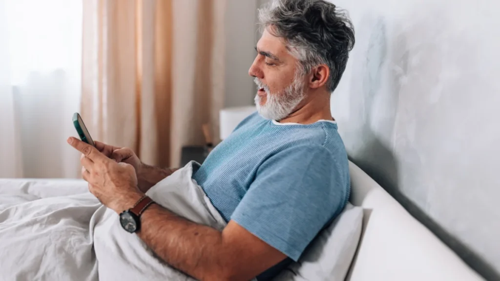 Mature man yawning while reviewing his smartphone sleep log to investigate why he continues to wake up tired after 50.