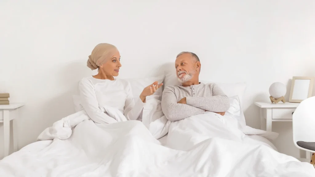 Mature woman in a head wrap gestures while talking to her partner in bed to discuss serious nighttime breathing concerns.