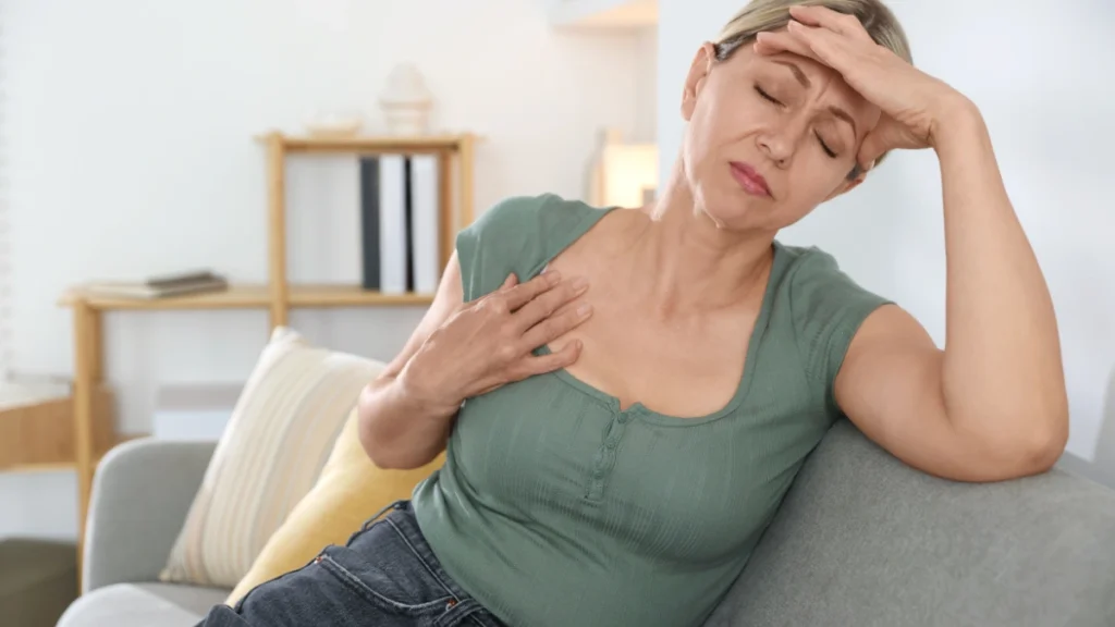 Mature woman holding her chest on a grey sofa to recognize dangerous fatigue red flags.