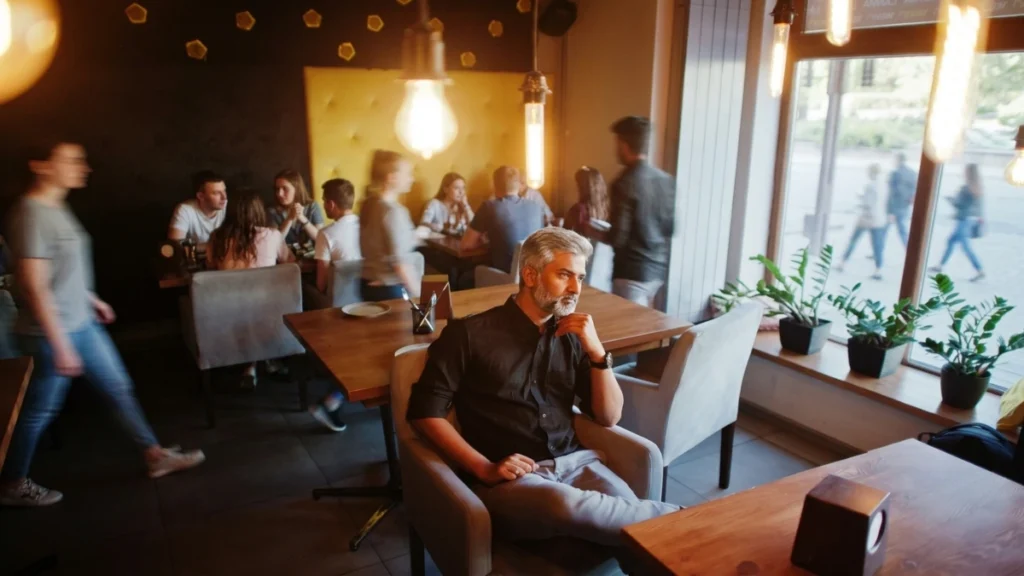 Silver-haired man sitting with his hand on his chin in a bustling restaurant, managing the mental fatigue of untreated hearing loss.