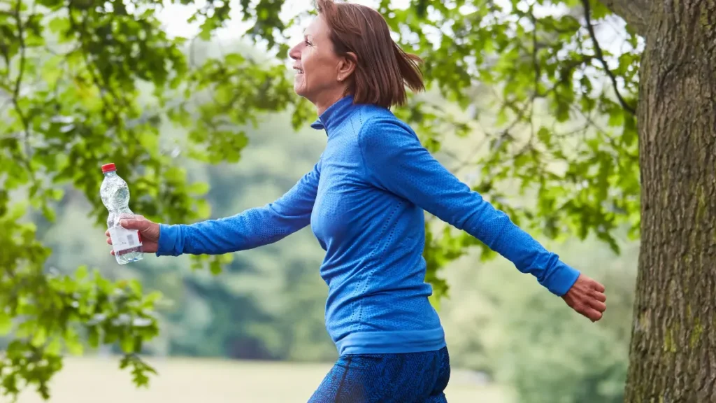 Mature woman power walking in a bright blue athletic top through a park to counter biological age acceleration.