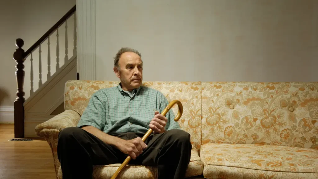 Older man sitting alone on a couch at home, holding a cane with a quiet, exhausted expression in a dim living room.