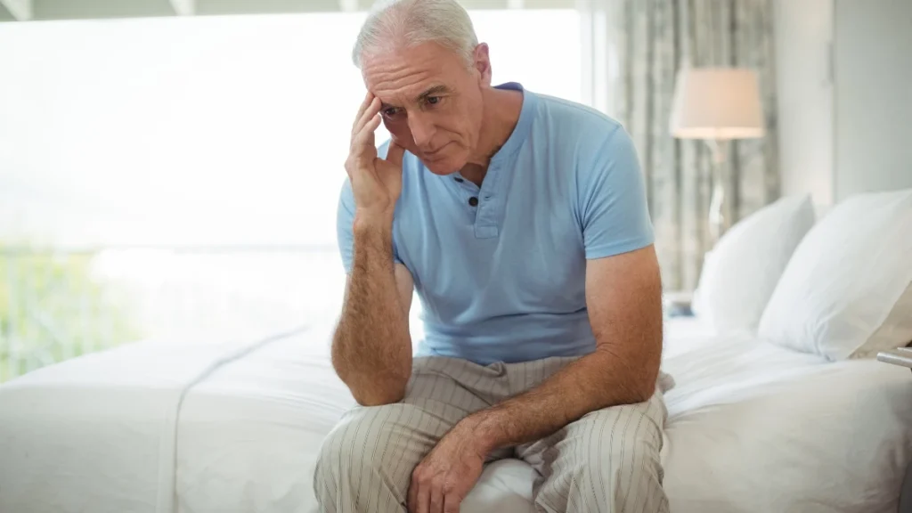 Mature man resting on bed edge with hand to temple to depict the physical burnout and cortisol strain of unresolved grief.