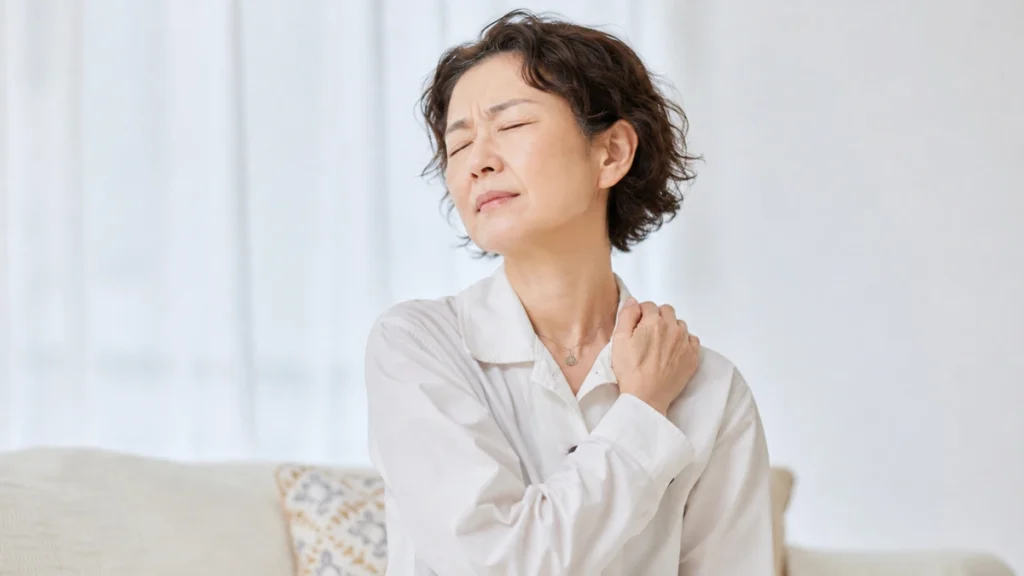 Mature woman rubbing a stiff, aching shoulder while seated on the sofa in early morning.