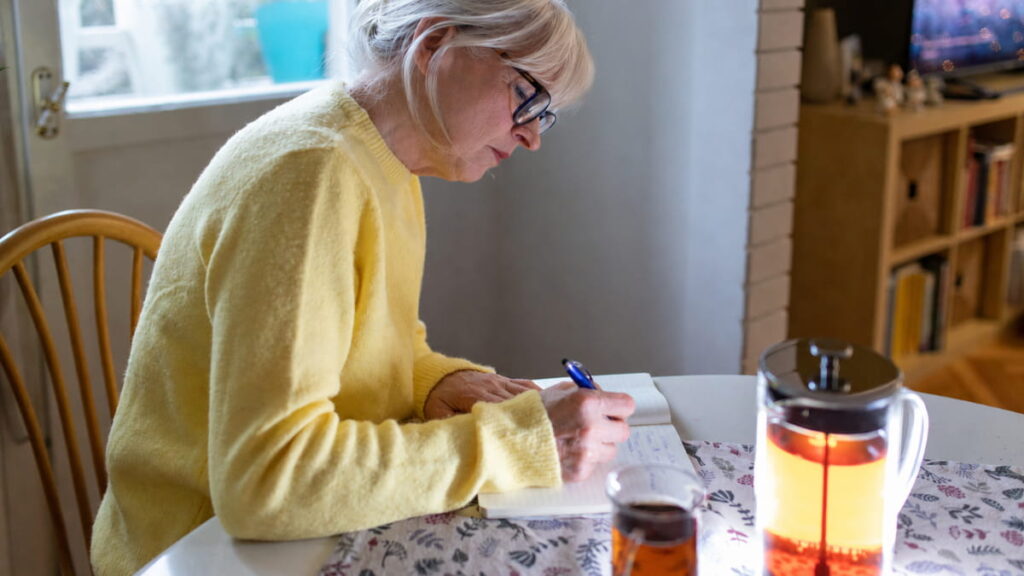 Woman in her 60s writing in a notebook at a kitchen table, recording daily symptoms and energy levels to bring to her next doctor's appointment.
