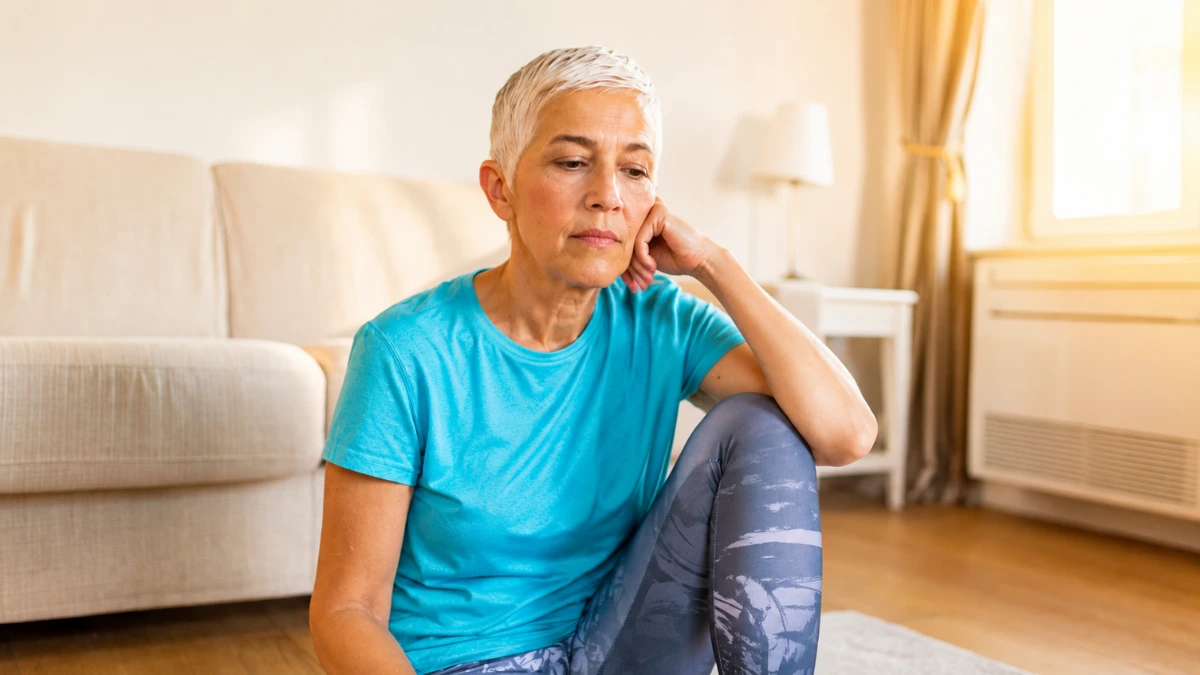 Woman in workout clothes sitting still on living room floor, expression flat and heavy after exercise.