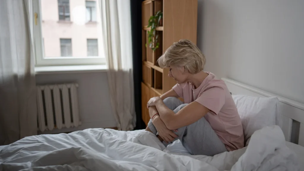 Woman in her late fifties seated on bed in morning light, knees drawn up, expression heavy and unrested after exercise.