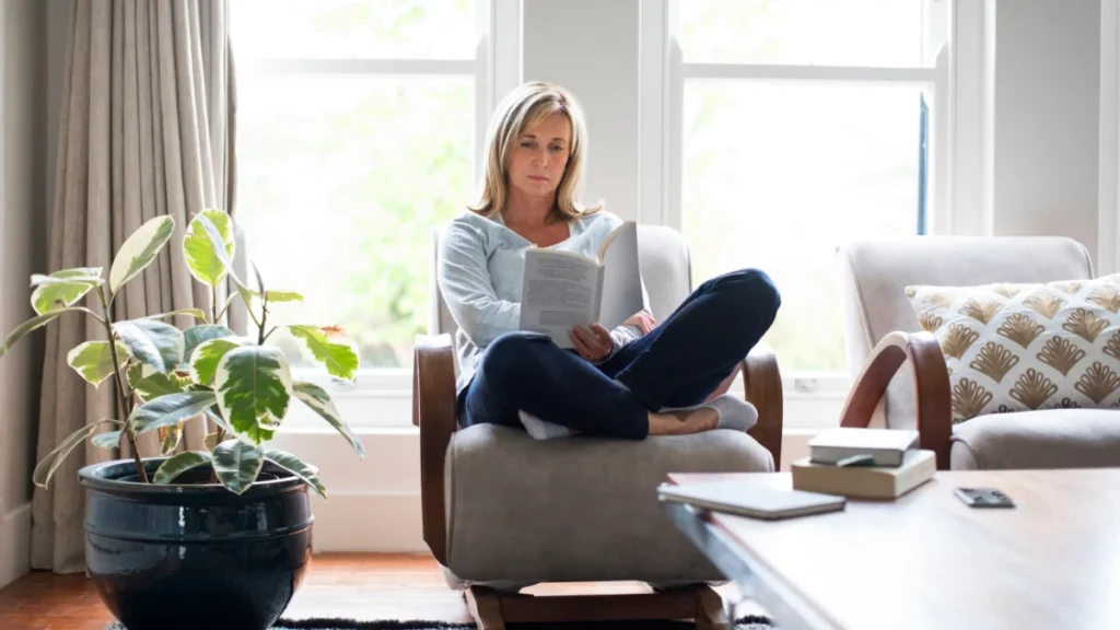Woman seated in reading chair near sunlit window with book open, expression calm and settled, embodying purposeful rest.