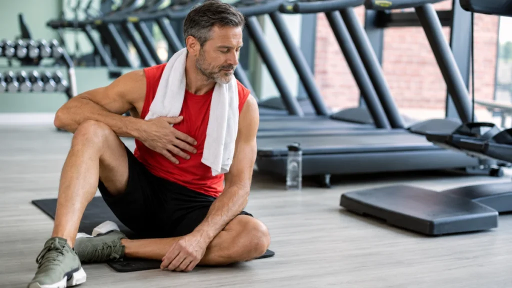 Man sitting on a gym floor after a workout, hand resting on his chest, eyes closed in recovery.