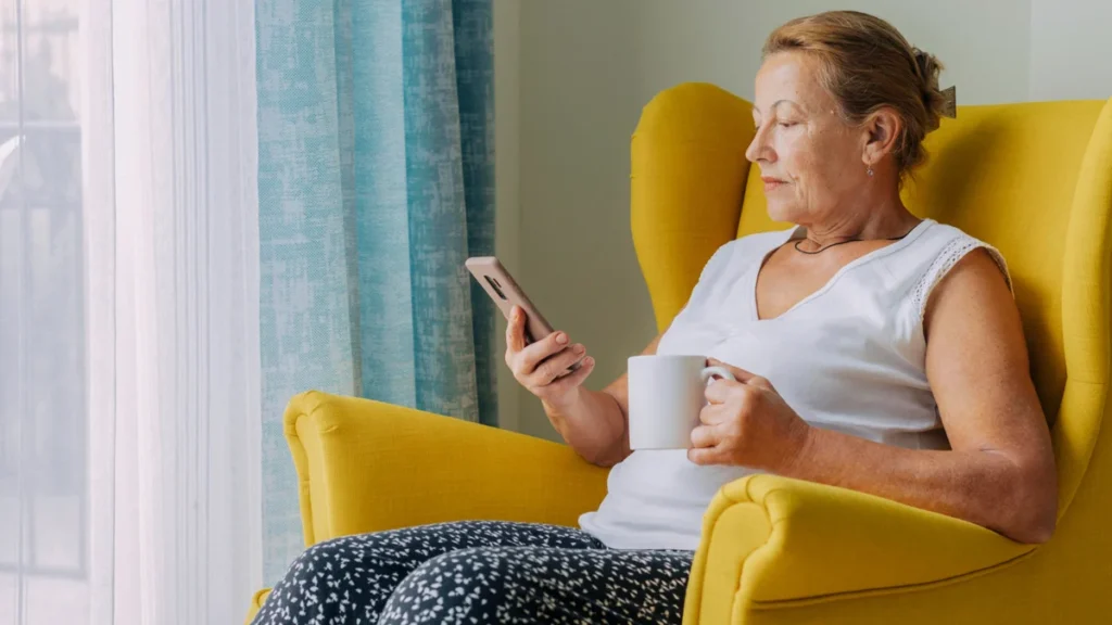 Woman in her 60s sitting upright at home reviewing her phone with calm focus to contact her doctor.