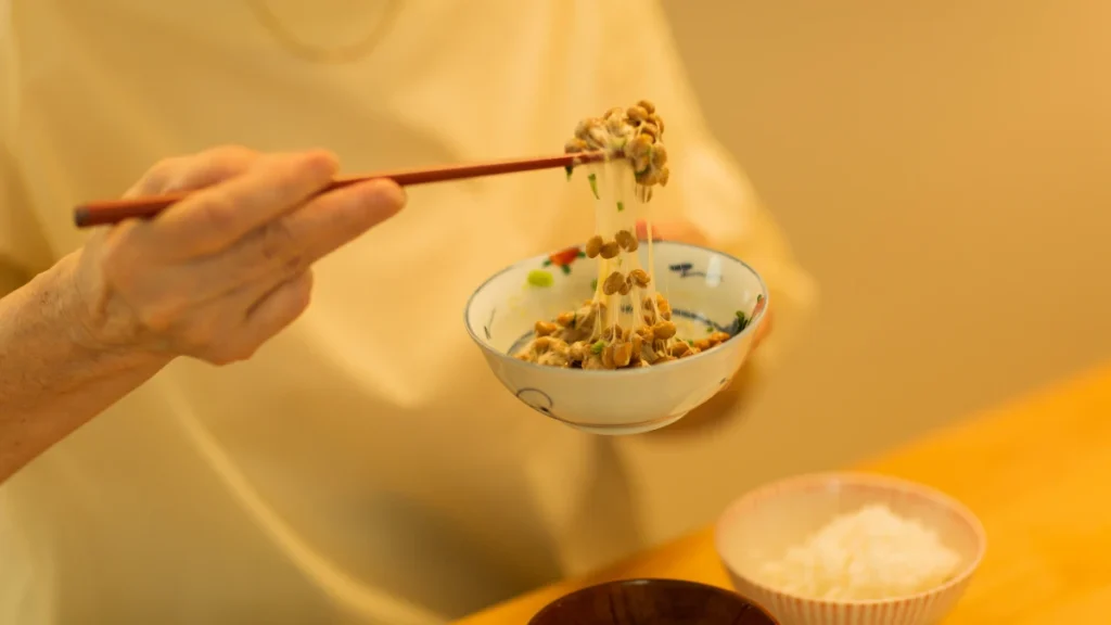 Woman in 60s eating natto with chopsticks at kitchen table, fermented soybeans as part of daily bone health routine.