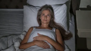 Woman in her 50s lying awake in a dark bedroom, eyes open, waiting for nervous system wind-down that hasn't arrived.