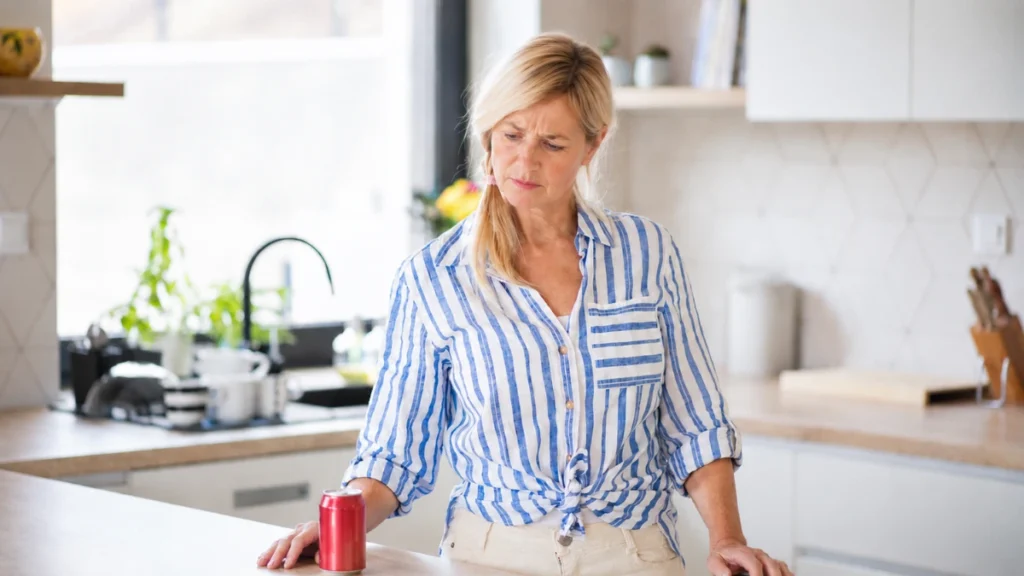 Woman in her late 50s pausing at a kitchen counter beside a soda can with a concerned expression after checking for hidden sweeteners.