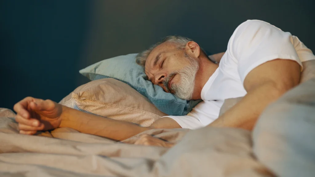 Man in his 50s lying in a warmly lit bedroom, hand on a cooling mattress pad, supporting deep sleep memory