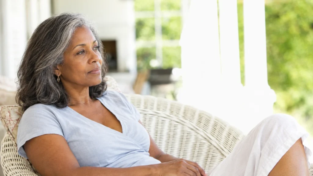 Woman in her 60s seated in warm morning light, expression quietly reflective, absorbing the slow toll of years of disrupted weekend sleep.