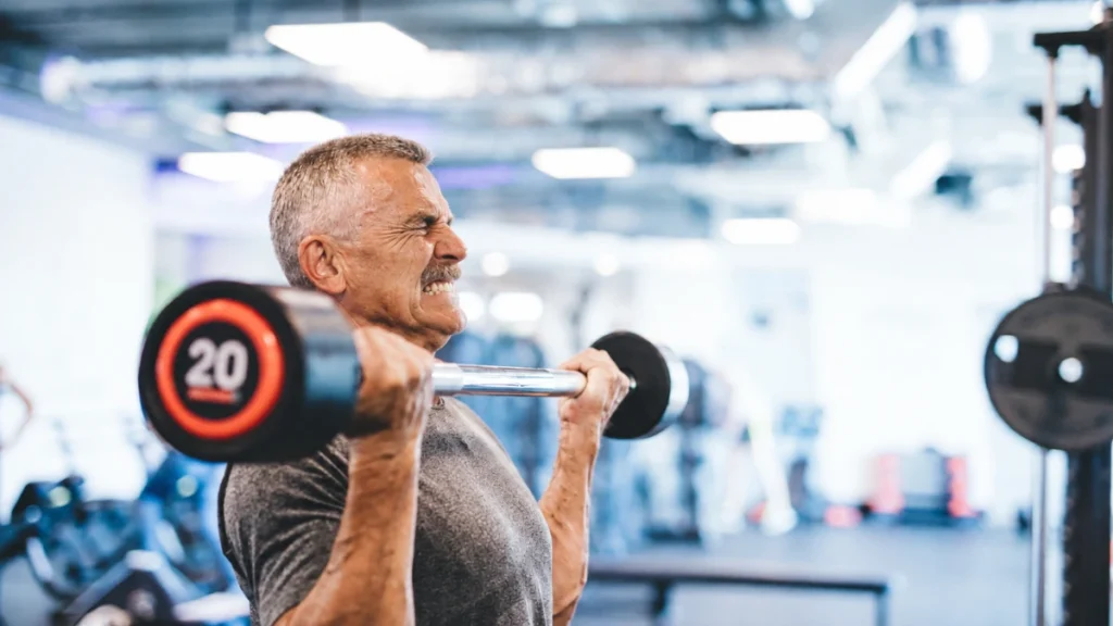 Man in his 50s grimacing through a moderate barbell curl, showing signs of nervous system fatigue that makes manageable weights feel impossible.