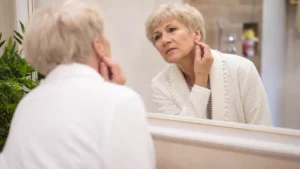 Woman in her 60s examining her earlobe in a mirror, checking for heart after 60 warning signs visible on the skin.