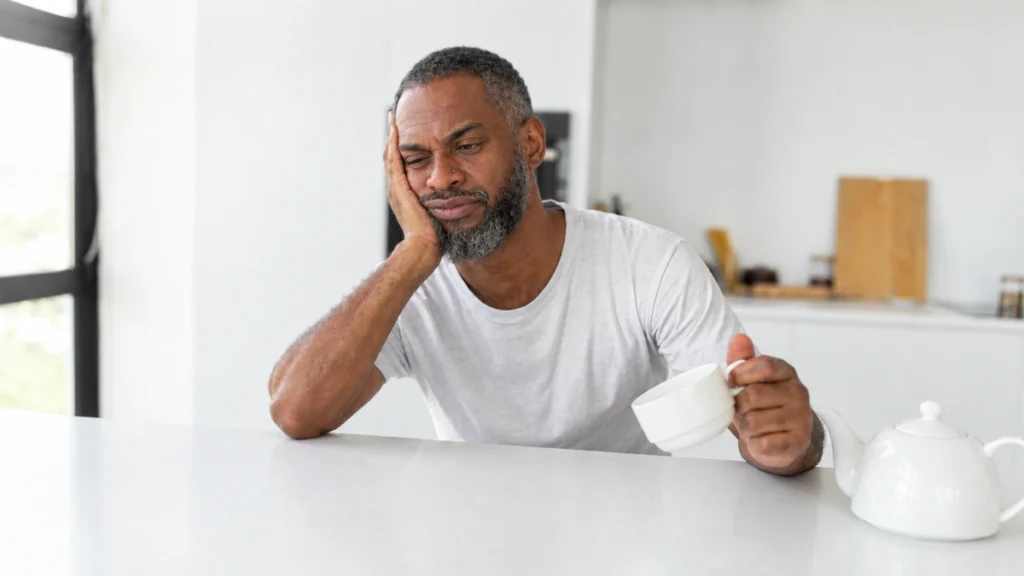 Tired man in his 50s sitting at a kitchen counter holding a coffee cup, head resting on his hand after a sleepless night.