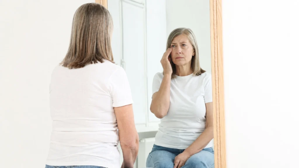 Woman in her 50s touching the corner of her eye in a mirror reflection, expression showing concern after noticing an unexpected eyelid flutter.