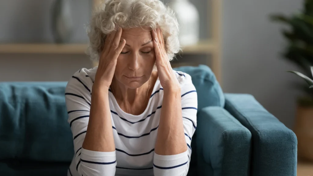 Woman in her 60s sitting on a sofa with both hands pressed to her temples and eyes closed, carrying the familiar weight of a recurring headache.
