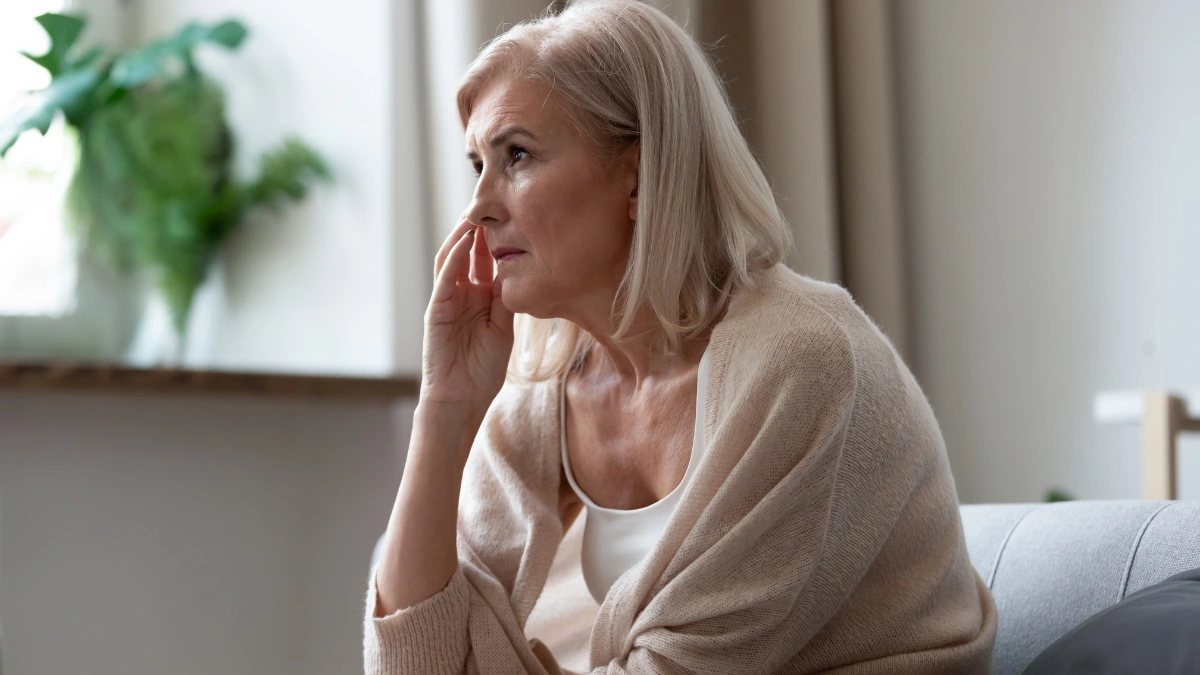 Older woman on living room couch hand raised to face pausing mid-thought as brain losing volume dims recall.